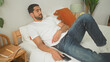 © Krakenimages.com - Man holds smartphone with hand on stomach, legs bent, reclined on bed in bedroom with notebooks and lamp visible; contemplation.