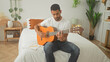© Krakenimages.com - Man playing an acoustic guitar on a bed with hands fretting strings in a studio space, jotting lyrics in a notebook beside him; songwriting calm.