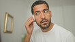 © Krakenimages.com - Man dabs a cotton pad to his eye and wipes cheek while inspecting skin at a vanity mirror inside a building; calm grooming.