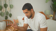 © Krakenimages.com - Man sits on bed gesturing with hand and smiling, wearing white tshirt near plant and notebook on bedside in building bedroom; calm reflection.