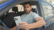 © Krakenimages.com - Man in driver seat of blue car holding smartphone with right hand and arm out the window, checking messages on screen on street; quiet concentration.