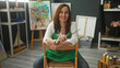 © Krakenimages.com - Woman artist in green apron sits on a chair backward, arms resting on the back while holding a paintbrush and smiling amid easels and paint bottles in studio; creative calm.