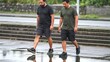 © Sheikhah - Two men walk over puddled pavement with steady strides and serious expressions. Outdoor walking in damp street setting feels calm and practical, with reflected legs visible in water.