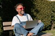 © Andrii Zastrozhnov - Happy mature man in denim overalls and red sunglasses working on a laptop on a park bench for outdoor remote work and lifestyle banner