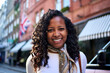 © CarlosBarquero - Cheerful millennial woman holds her backpack straps looking at camera on a lively London street, with Union Jack and striped awnings behind her, capturing a bright travel moment with confident energy