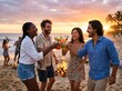 © Glam - Friends celebrating at beach bonfire with colorful drinks during sunset