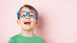 © SerPak - Young boy smiling and laughing while wearing bright blue eyeglasses against a soft pink background. Cheerful childhood portrait with copy space.