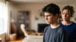 © Natali - A boy stands in a living room with an angry expression. He looks upset while a woman stands behind him, showing concern. The room has furniture and