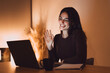 © Daniel - Young woman smiling and waving at her laptop screen during a video call, communicating remotely from a home office at night, showcasing modern virtual connection and remote work lifestyle