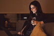 © Daniel - Woman relaxing on a sofa at night, smiling while using a laptop and holding a glass of red wine, embodying a cozy and personal leisure lifestyle at home