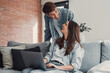 © Daniel - Happy young couple enjoying time together at home, a woman sitting on the couch using a laptop and a man standing behind her, both smiling and looking at each other