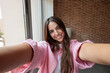 © Daniel - Young woman smiling broadly, stretching arms out, and capturing a selfie indoors, showing a happy and confident lifestyle moment in natural light against a brick wall and window