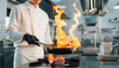 © macashop - A professional chef in a white jacket flambés food in a busy commercial kitchen, tossing a flaming pan over a gas stove to create dramatic flames and dynamic culinary action.