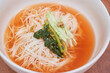 © 진영김 - Korean summer noodle dish yeolmu guksu, thin wheat noodles in cold spicy red broth, topped with fermented radish green kimchi, julienned cucumber, and mustard sauce, on a wooden table.