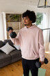 © Wavebreak Media - Man wearing pink hoodie lifting black dumbbells curling left arm on green mat in living room