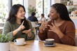 © Jafree - Two smiling women talking in sign language at cafe table with coffee cups. Asian and Black female friends communicating with hand gestures. Deaf culture and nonverbal communication concept