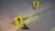 © Dark Artistic - Crime scene investigators placing yellow evidence markers next to ammunition cartridges and tape on a dusty concrete floor, documenting forensic evidence for legal investigation