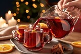 Hot mulled wine being poured from glass pitcher into clear mugs with orange slices, cinnamon sticks and star anise on wooden tray, warm candlelight background