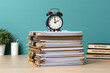 © Akash - A black alarm clock sitting on top of a huge stack of paperwork on a wooden desk. Useful for business deadline and time management concepts.