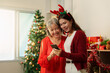 © Natee Meepian - Asian mother and adult daughter using smartphone near Christmas tree for holiday connection