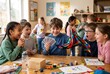 © Kassiopeia  - A group of children plays together at a table in a classroom. Some are showing excitement while one child holds colorful fabric and playing cards. They are enjoying their time after school