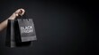 © MSTMAYA - A hand holding two black shopping bags with the text black friday isolated against a dark background, for retail sales, and consumer shopping events