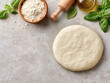 © Liliya - Fresh pizza dough on a kitchen table with flour, basil, olive oil, and a rolling pin, ready for homemade baking, top view.