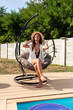 © milanmarkovic78 - A woman enjoys a leisurely moment in a stylish hanging chair near a pool, surrounded by greenery. She smiles while wearing a hat and casual attire, embodying a relaxed, joyful atmosphere in bright sun