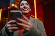 © BullRun - Young multiracial lady with smartphone looking away and smiling in city at night, mobile connectivity, digital lifestyle, 5G wireless network, startup culture, inclusion, urban traveler mood.