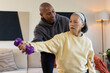 © Wavebreak Media - Senior Asian woman being guided by African American man lifting purple dumbbell on sofa at home