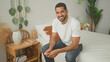 © Krakenimages.com - Man sitting on bed holding smartphone and smiling, wearing white tee and jeans beside rattan nightstand with lamp, notebook and plants in bedroom; relaxed ease.