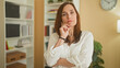 © Krakenimages.com - Woman touching chin with crossed arms in building office interior with bookshelf, clock and plant visible; contemplation.