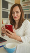 © Krakenimages.com - Middle age woman holding red smartphone, finger tapping screen while seated at desk in office building amid bookshelves; quiet concentration calm.