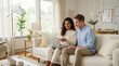 © mashimara - A happy couple sitting on a sofa in a bright living room while browsing online on a laptop