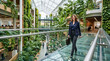 © MOON - confident professional businesswoman wearing navy suit and holding briefcase walking along transparent glass skybridge inside eco friendly modern corporate atrium with lush living walls