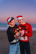 © Renata Hamuda - Two male friends wearing ugly Christmas sweaters and Santa hats holding sparklers on the beach