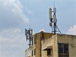 © yiam - Weathered building rooftop hosts two distinct telecommunications antenna arrays against bright blue sky with scattered clouds
