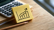 © Denis Yevtekhov - Yellow sticky note carrying a simple rising business growth chart beside a white calculator quarterly ledger and a sharpened pencil aligned beneath the note light oak desk with