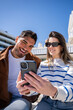 © CarlosBarquero - Vertical. Young couple sit outdoors and smile while looking at a smartphone together in a modern residential area, sharing digital content during leisure time.