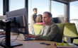 © CarlosBarquero - Focused mature businessman typing on a laptop at his desk while colleagues work in the background. Bright corporate scene conveying concentration, responsibility and productivity.