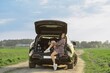 © Aleksei Isachenko/imageBROKER - A woman in casual attire leans against the trunk of her car, enjoying the sunny weather on a dirt country road. Lush green fields stretch out in the background, showcasing a peaceful rural landscape