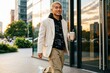 © Leonardo - Un joven empresario relajado saliendo de un edificio de oficinas acristalado durante la hora dorada, vistiendo una americana color crema de corte amplio sobre una sudadera