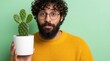 © Vadym - Young bearded man holds small cactus in white pot. Funny guy looks surprised at succulent plant with green prickly leaves. Man wearing yellow sweater poses with indoor houseplant against light green