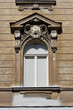 © nahhan - A detailed front view captures an arched white window on an old brown building facade featuring weathered baroque ornaments and a triangular pediment.