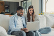 © Daniel - Couple uses a laptop together on a sofa at home, smiling as they browse online. Black man and Caucasian woman share technology in a bright living room with an open kitchen, enjoying connected life.