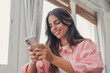 © Daniel - Young woman uses a smartphone for messaging at home, smiling while typing on her mobile device near a window. Bright natural light supports everyday communication, internet and social media use.