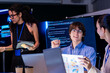 © ultramansk - A curly haired analyst and a programmer at his laptop explains the next task at his laptop in a dark technology office lit in violet and blue, an image of mentoring in technology