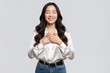 © Maryna - Smiling young Asian woman places hands on chest with closed eyes. Grateful female feels relief, peace in studio. Happy lady wearing white silk blouse, blue jeans shows mindful emotion, gratitude.