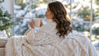© Nattapol - Woman holding ceramic mug wrapped in oversized knit blanket enjoying cozy winter comfort indoors
