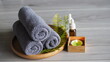 © kanjana - Spa and wellness still life featuring neatly rolled gray towels, aromatherapy essential oil bottles, fresh green fern leaves, and a glowing candle arranged on a wooden tray. The calming natural compos
